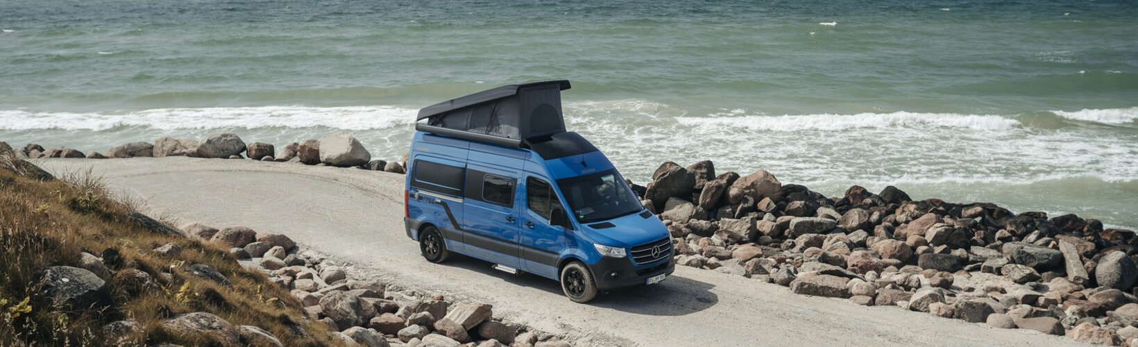 Blauer Hymer Camper Van mit Hochdach fährt an Küste mit Blick aufs Meer