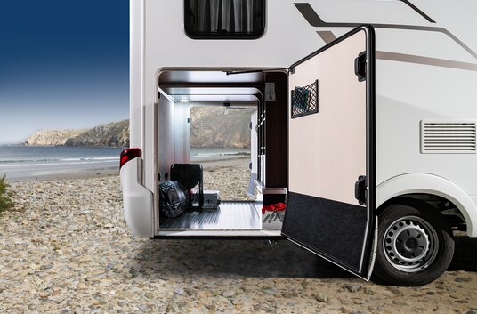 The rear garage of the HYMER motorhome, open on both sides and loaded with luggage, is on the beach with a blue sky and rocky cliffs