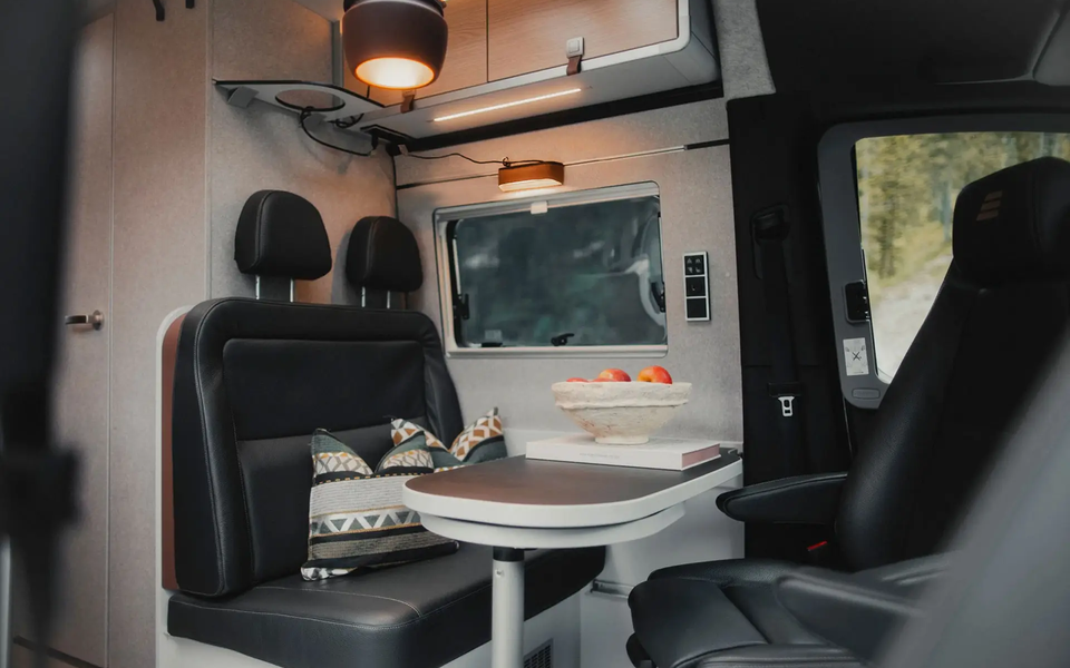 Wohnraum in einem modernen Camper Van, gedeckter Tisch mit Sitzgruppe mit dunklem Leder, die beiden Fahrersitze sind zum Tisch gedreht, oberhalb der Sitzgruppe sind Dachstauschränken und eine braune Pendelleuchte