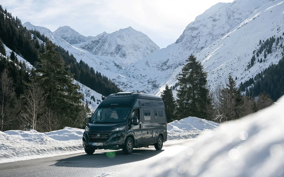 Dunkler HYMER Campervan, der durch eine bewaldete, winterliche Landschaft mit schneebedeckten Bergen im Hintergrund fährt.	