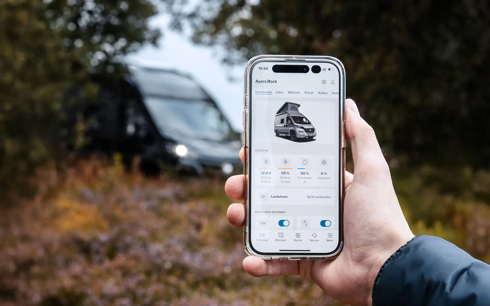 Hand, die ein Handy hält, auf dem der Inhalt einer App zur Fahrzeugsteuerung eines Campervans zu sehen ist. Im Hintergrund ist unscharf ein Campervan zu sehen, der in einer bewaldeten Umgebung steht.	