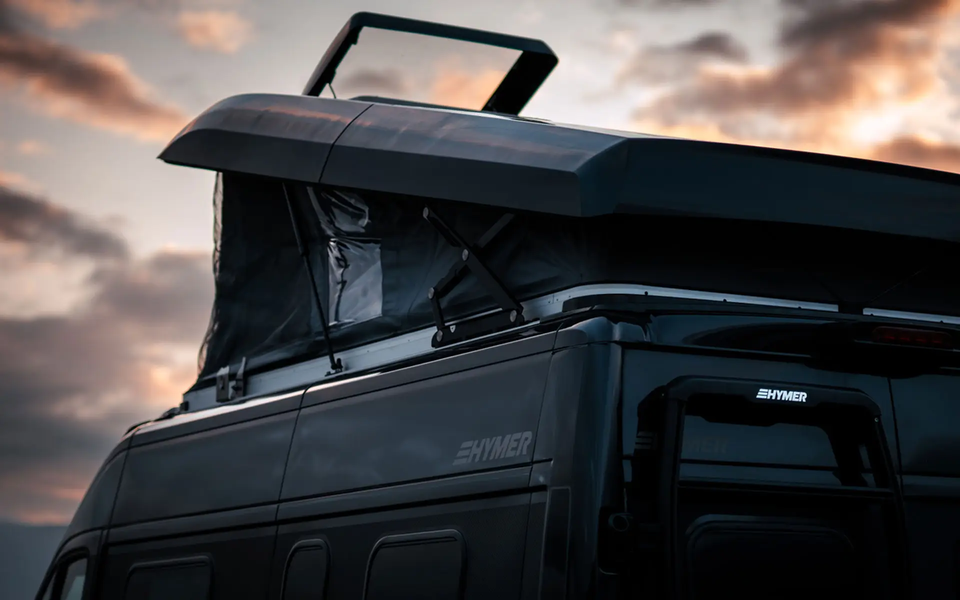 Detailansicht eines schwarzen, aufgestellten Schlafdachs bei einem schwarzen HYMER Campervan vor einem dämmerigen Abendhimmel. Das Schlafdach ist von hinten seitlich zu sehen; man kann den Stoßdämpfer und den Scherenmechanismus sehen, der das Schlafdach aufhält.	