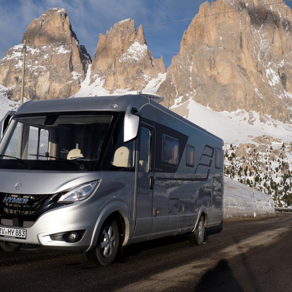 Ein silberner HYMER B-Klasse MasterLine I steht bei sonnigem Wetter auf einer asphaltierten Straße, im Hintergrund sind Berge und Schnee zu sehen. 