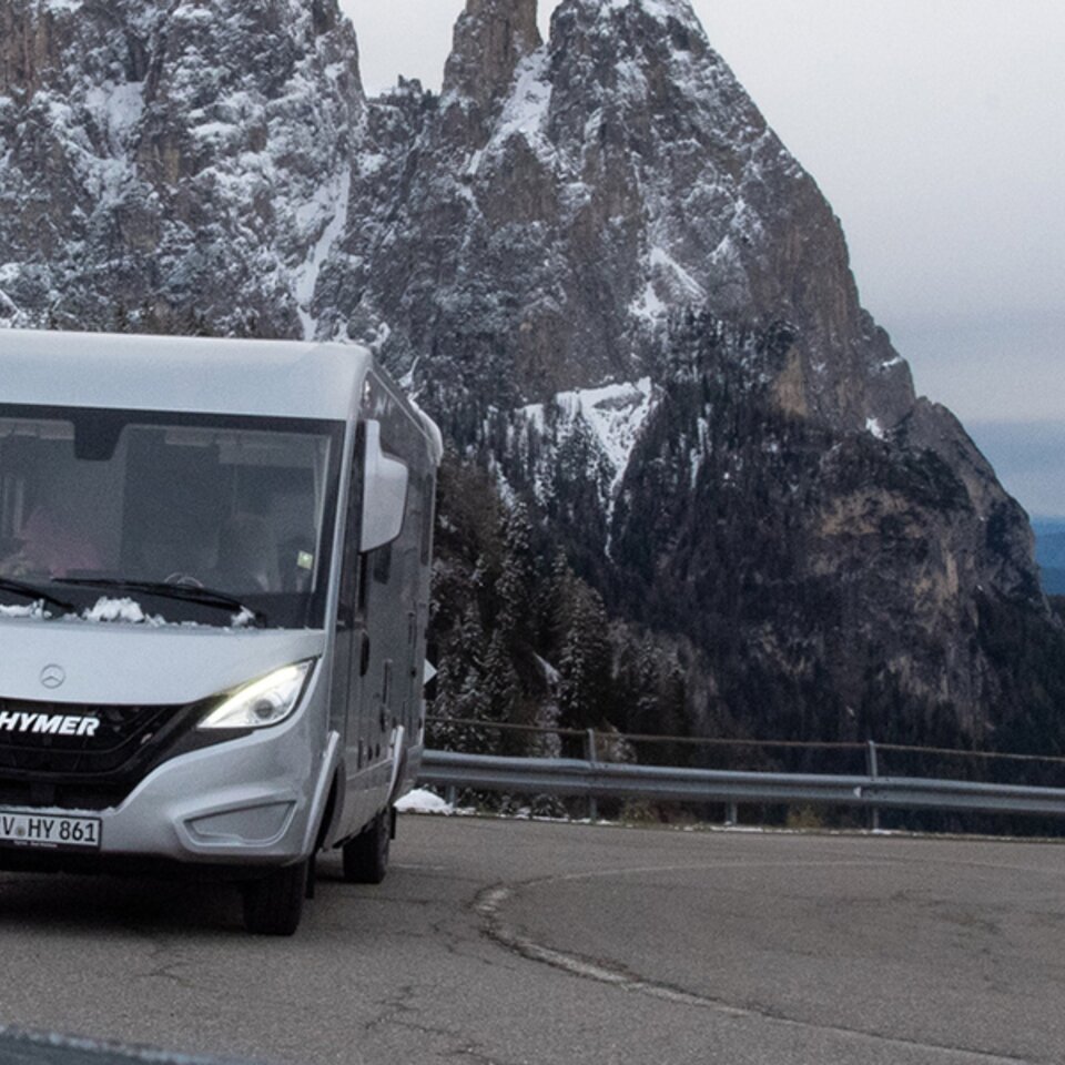Ein grauer HYMER B-Klasse ModernComfort I steht mit leuchtenden Scheinwerfern in der Kurve einer Gebirgsstraße. Auf den Bergen dahinter liegt Schnee.
