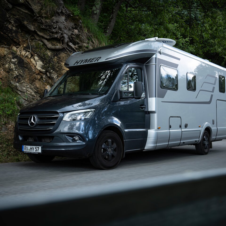Der HYMER B-Klasse MasterLine T mit dunklem Fahrerhaus und grauem Aufbau steht auf einer Straße mit felsiger Böschung und grünen Bäumen am Rand.