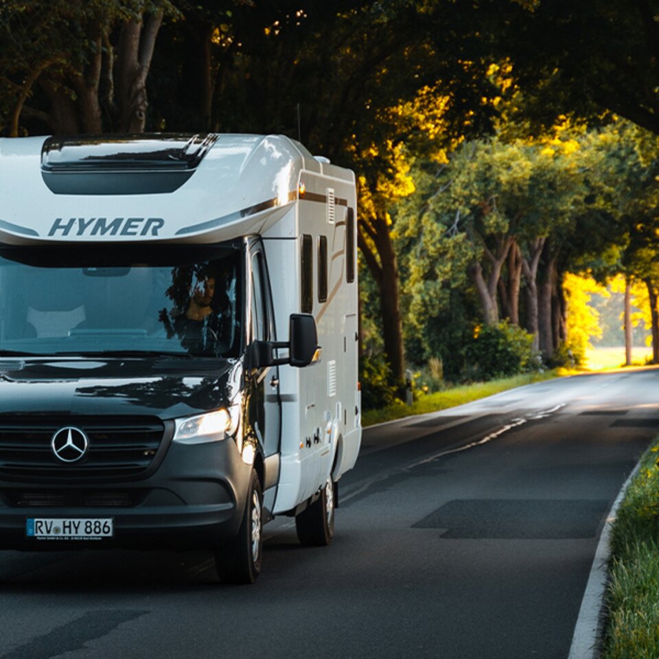 Ein schwarz-weißer HYMER B-Klasse ModernComfort T fährt bei sonnigem Wetter mit leuchtenden Scheinwerfern durch eine Allee mit grün belaubten Bäumen.