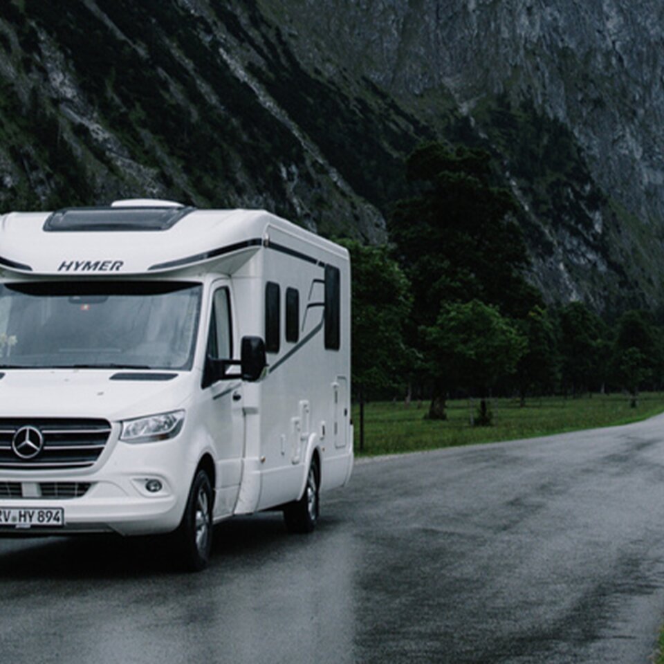 Ein weißer HYMER Tramp S steht auf einer schmalen, regennassen Straße in bergiger Landschaft, im Hintergrund sind Berge, Bäume und Wiesen zu sehen.