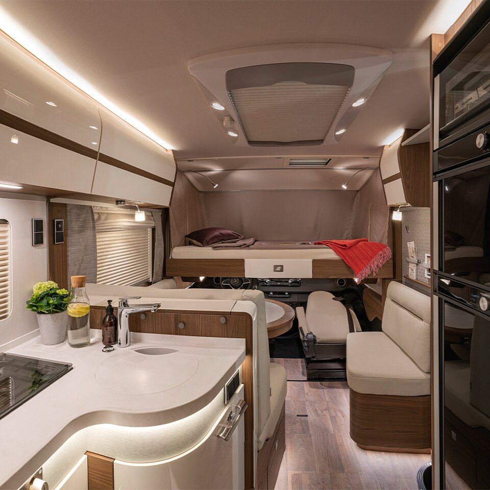 Blick in das Mercedes Wohnmobil B-Klasse MasterLine I mit Holzdekor, auf beleuchtete Küche, Sitzbank und Bett mit roter Decke und violettem Kissen. 