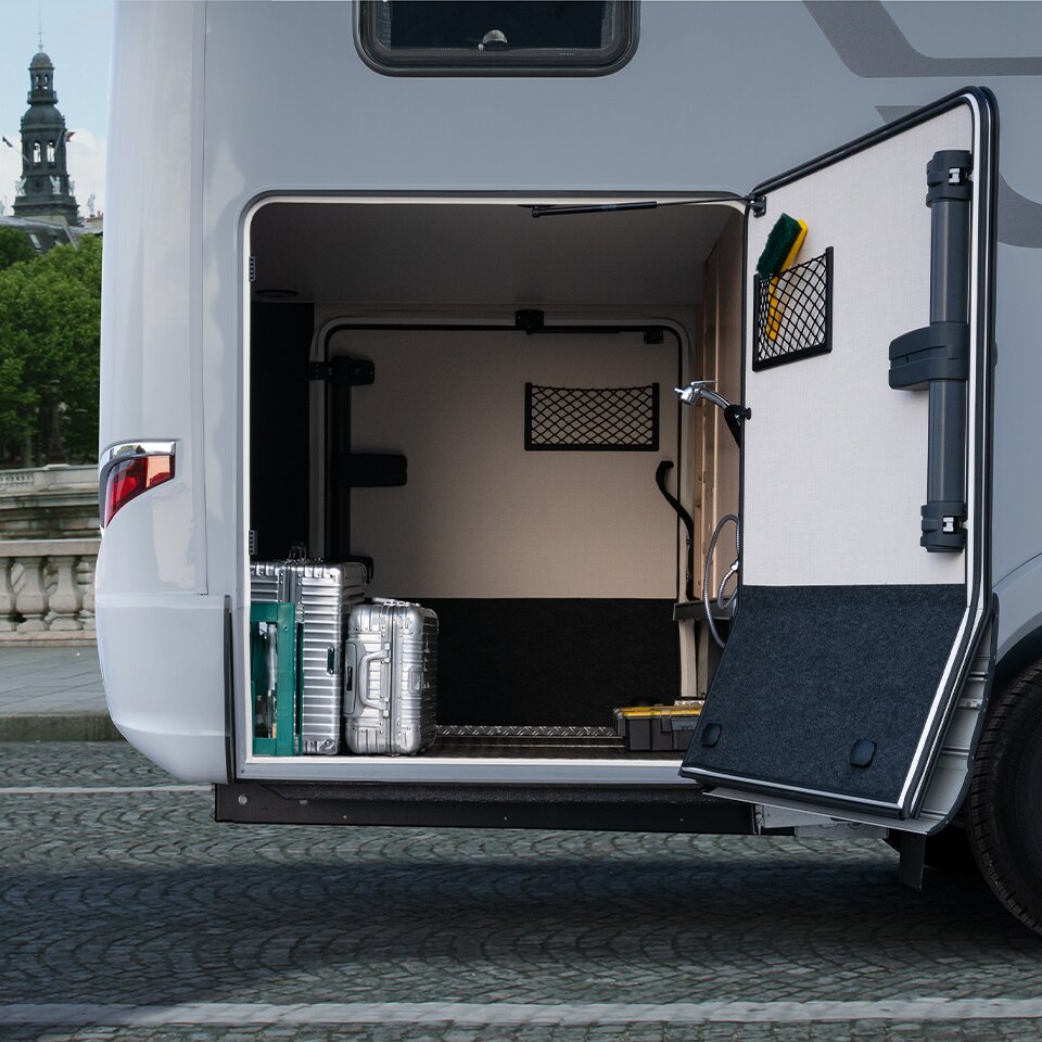 Eine Tür der Garage des HYMER B-Klasse MasterLine T ist geöffnet, es stehen silberne Koffer darin. Das Mercedes Wohnmobil steht auf grauem Pflaster.