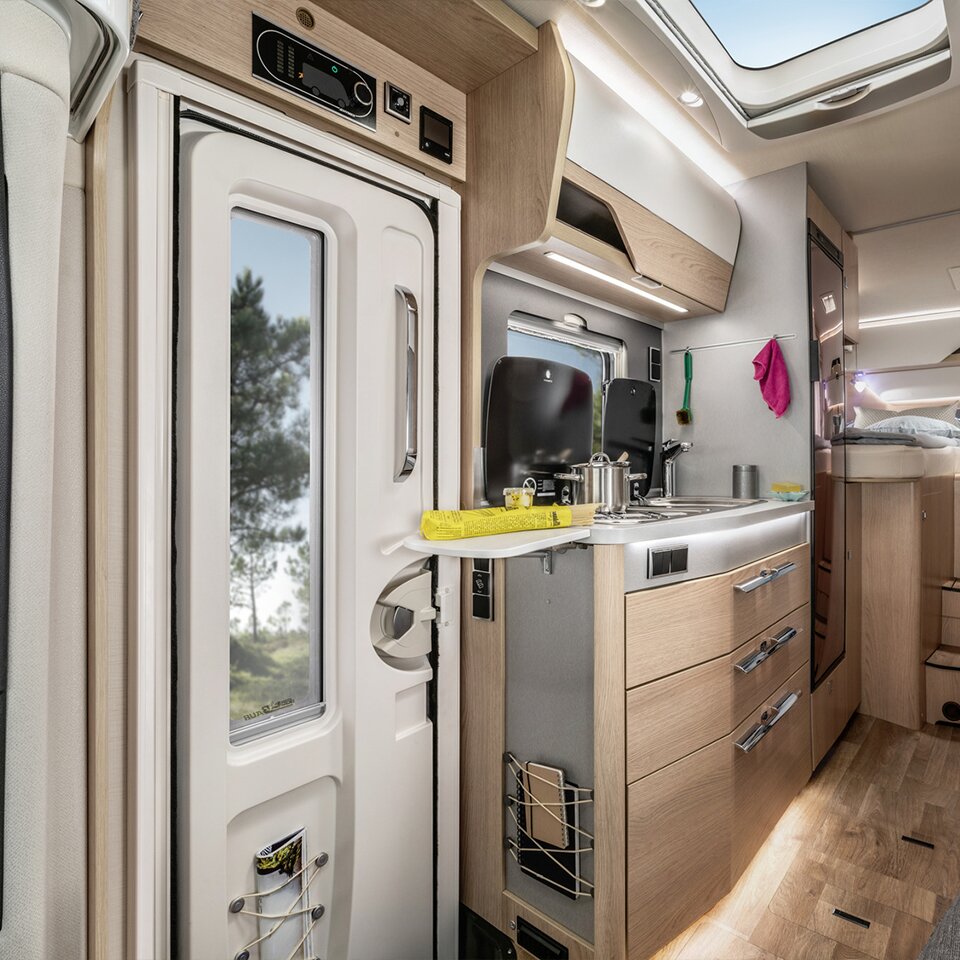 Blick auf den Kochbereich des HYMER B-Klasse Modern Comfort T in hellem Holzdekor, daneben die weiße Tür, durch deren Fenster Bäume zu sehen sind.