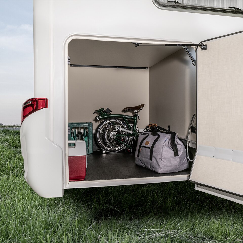 In der offenen Garage des HYMER Tramp S, einem Mercedes Sprinter Camper, stehen eine Reisetasche, ein Klapprad, ein Getränkekasten und Zubehör-Koffer.