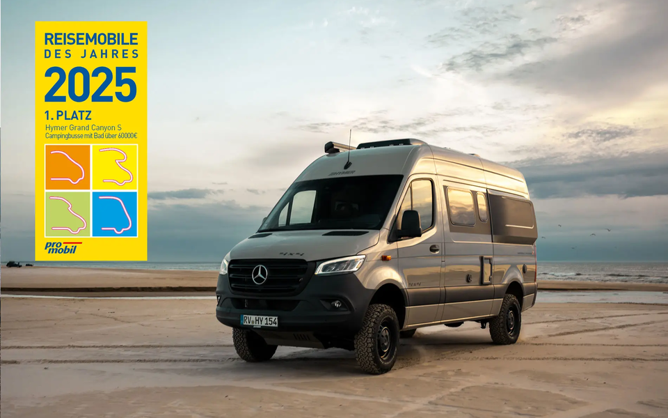 Vy från snett framifrån på en ljusgrå HYMER 4x4-campervan som står på en sandstrand. I bakgrunden syns havet och en lätt molnig himmel. Utmärkelselogotypen "Årets husbil 2025, 1:a plats Hymer Grand Canyon S" är placerad på bilden.	