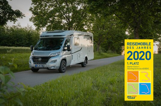 Wohnmobil HYMER Exsis-t fährt auf von grünen Wiesen und Bäumen gesäumter Straße / mit Auszeichnung Reisemobil des Jahres 2020