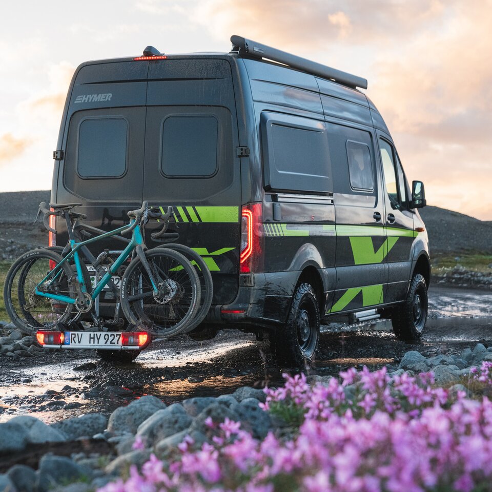 Il camper Sprinter 4x4 HYMER Grand Canyon S CrossOver è su un sentiero sterrato con fiori e erba, sul portabici ci sono biciclette.