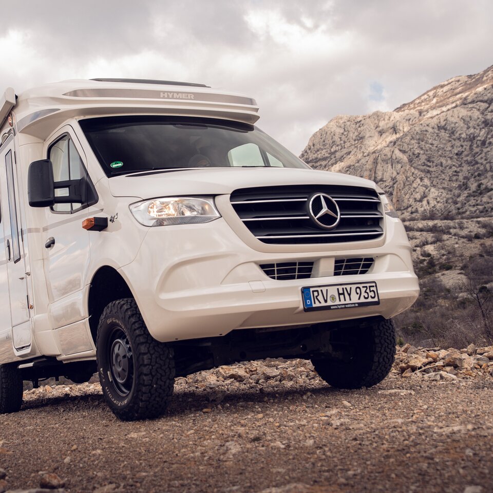 Il camper Mercedes Allrad HYMER ML-T è su una strada di pietra con colline rocciose e cielo leggermente nuvoloso sullo sfondo.