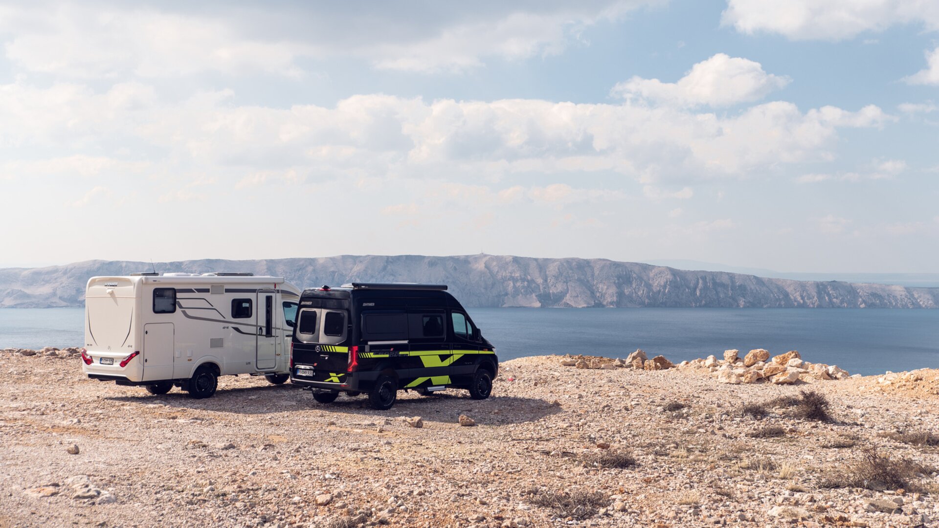 Due camper fuoristrada bianco e nero-verde sono su un piano soleggiato con vista sul mare, sullo sfondo ci sono scogliere.