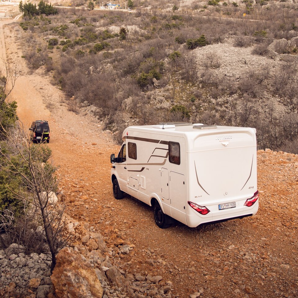 Due camper fuoristrada 4x4 di HYMER con motore Mercedes viaggiano su una strada collinosa attraverso un terreno roccioso e cespugli.