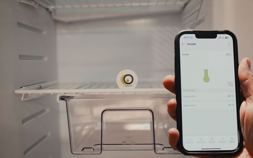 In the foreground, a hand holding a mobile phone displaying a temperature reading. In the background, the interior of an empty refrigerator with a sensor placed inside.	