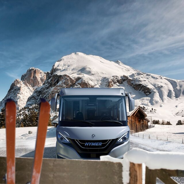 HYMER B-Class MasterLine in a sunny, alpine winter landscape with a wooden barn and a pair of skis leaning against a wooden fence