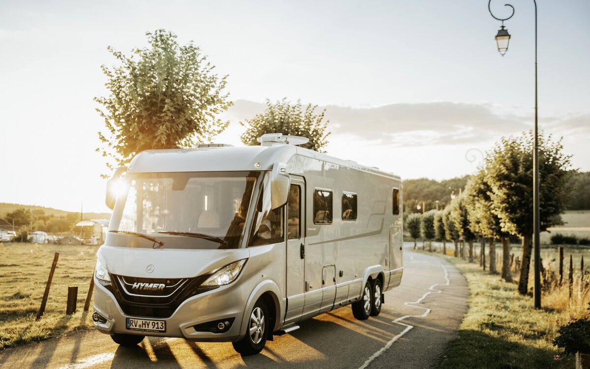 Double-axle HYMER B-Class MasterLine I drives along a tree-lined street in warm lighting