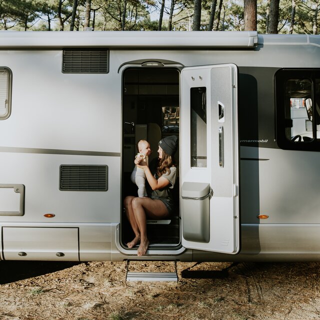 A young woman with a baby is sitting in the entrance area of a HYMER motorhome with the main door open and the rubbish bin integrated