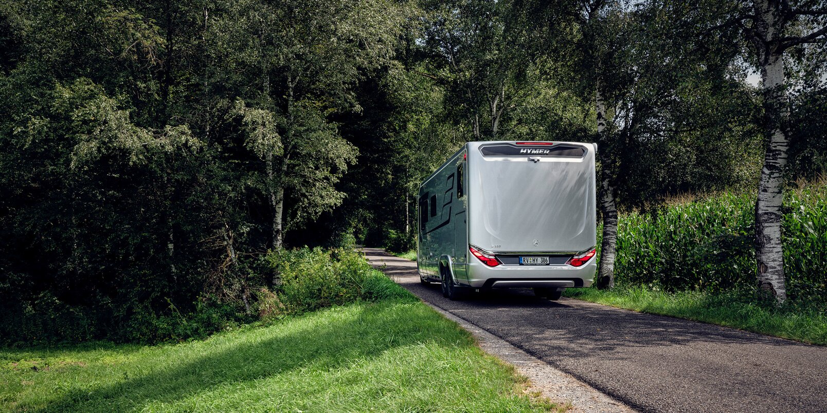 HYMER motorhome B-ML I 880 from behind on the road at the edge of the forest with a green meadow and maize field