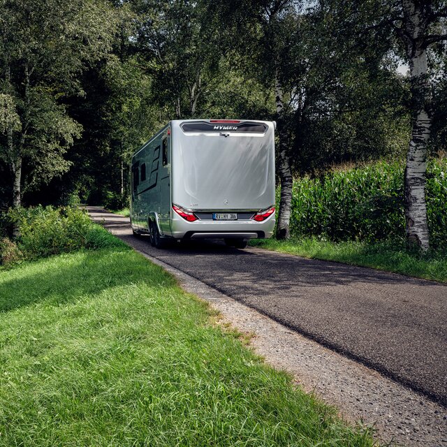 HYMER motorhome B-ML I 880 from behind on the road at the edge of the forest with a green meadow and maize field