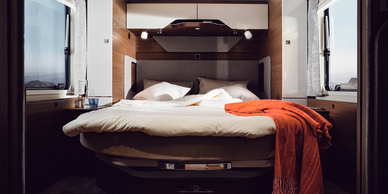 Queen-size bed with cream-colored bed linen and red decorative blanket in the rear of the HYMER B-ML I 890