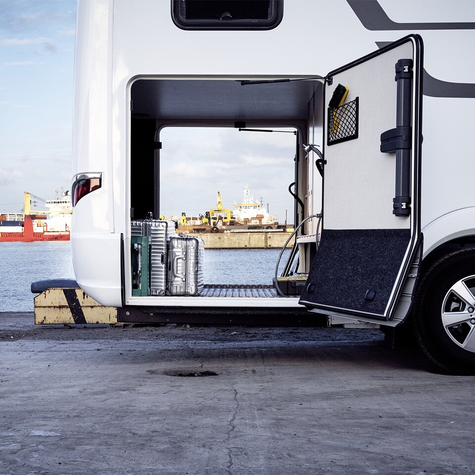 In der offenen Garage des HYMER B-Klasse Master Line I, einem Wohnmobil mit Mercedes-Triebkopf, stehen Koffer. Im Hintergrund ist ein Hafen zu sehen.