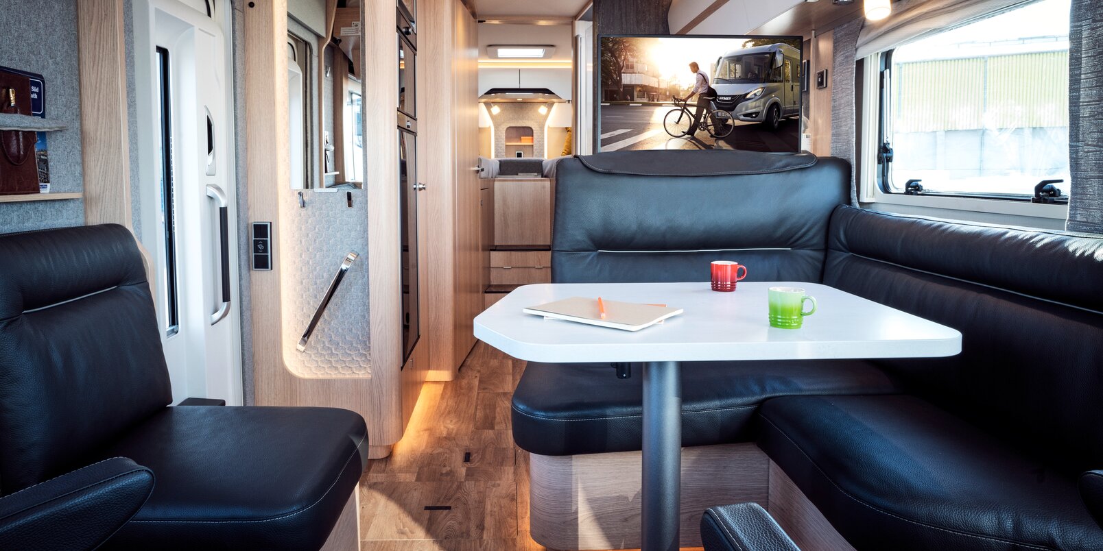 Seating group in black leather look with a laid table and a view of the TV plasma and rear area in the HYMER B MasterLine