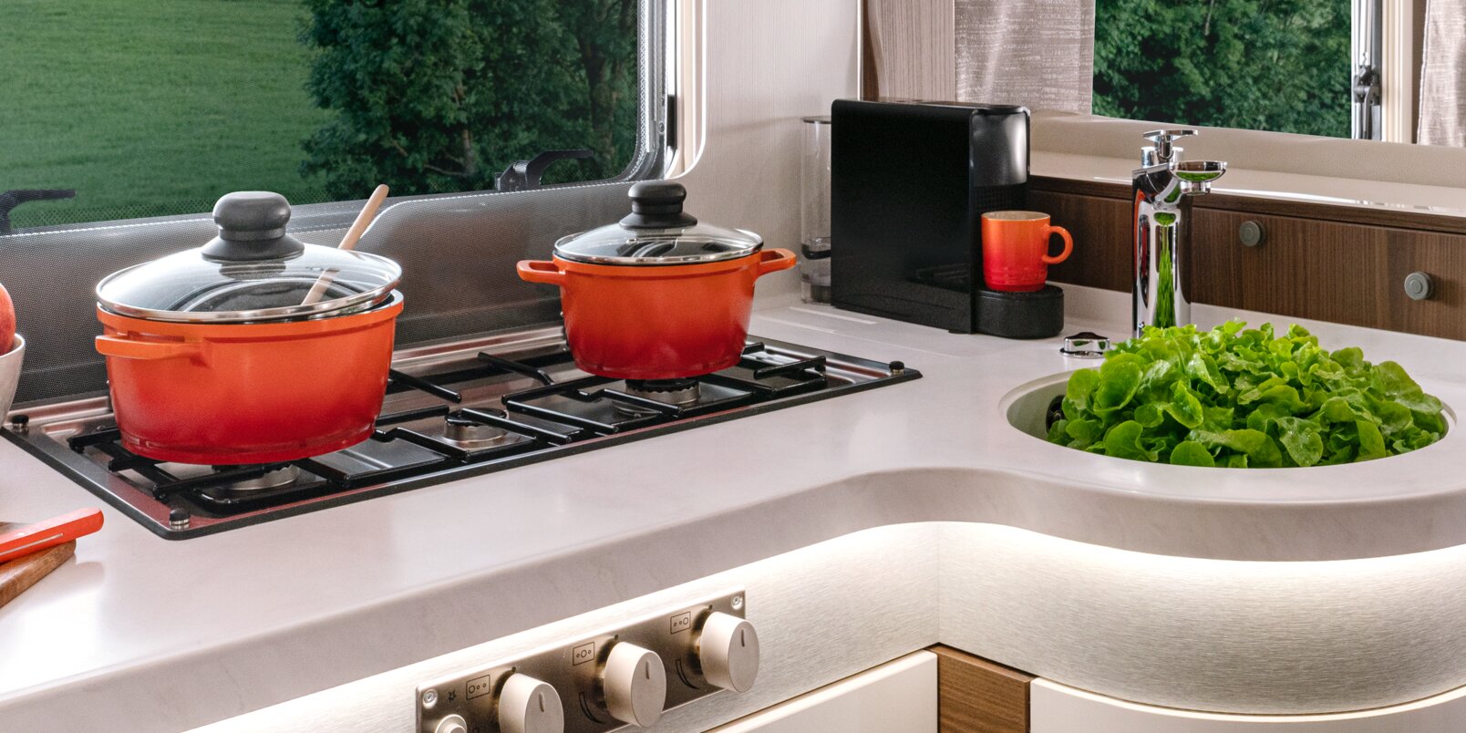Cucina con piano cottura e due pentole rosse, lavandino con dentro un’insalata e macchinetta del caffè nell’HYMER MasterLine