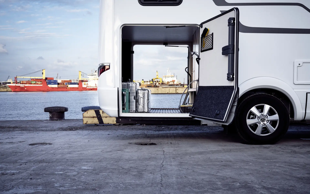 View through the rear garage of the HYMER B-ML T motorhome, loaded with silver suitcases, in front of the port facility with the red freighter