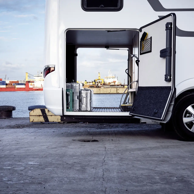 View through the rear garage of the HYMER B-ML T motorhome, loaded with silver suitcases, in front of the port facility with the red freighter