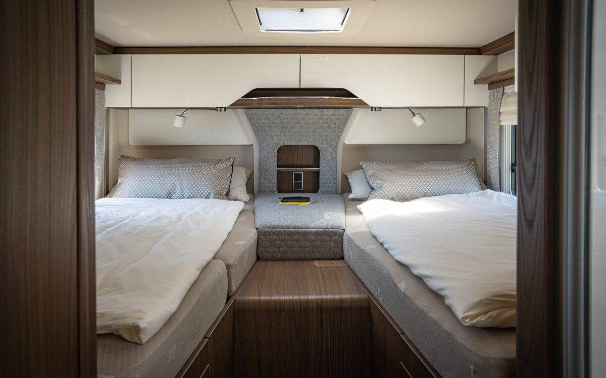 Sleeping area in the mobile home with two lengthways single beds, between them a center console for storage and for storing utensils and closed storage compartments above the head area