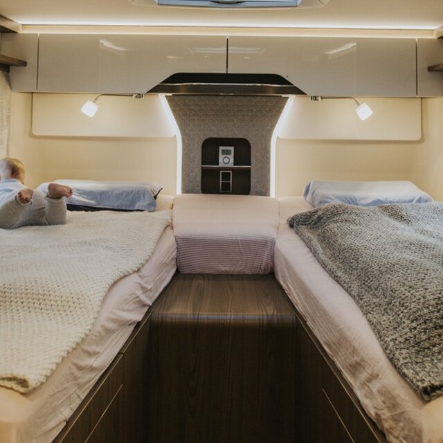 Toddler on left length single bed in rear of camper van; cozy living atmosphere thanks to pleasant ambient lighting