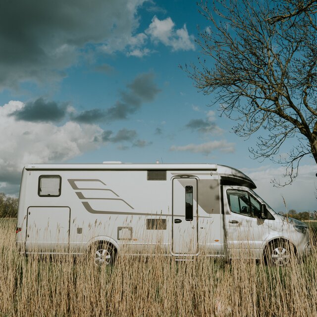 Side view of a HYMER motorhome under a partly cloudy sky in a rural area with grass in the foreground