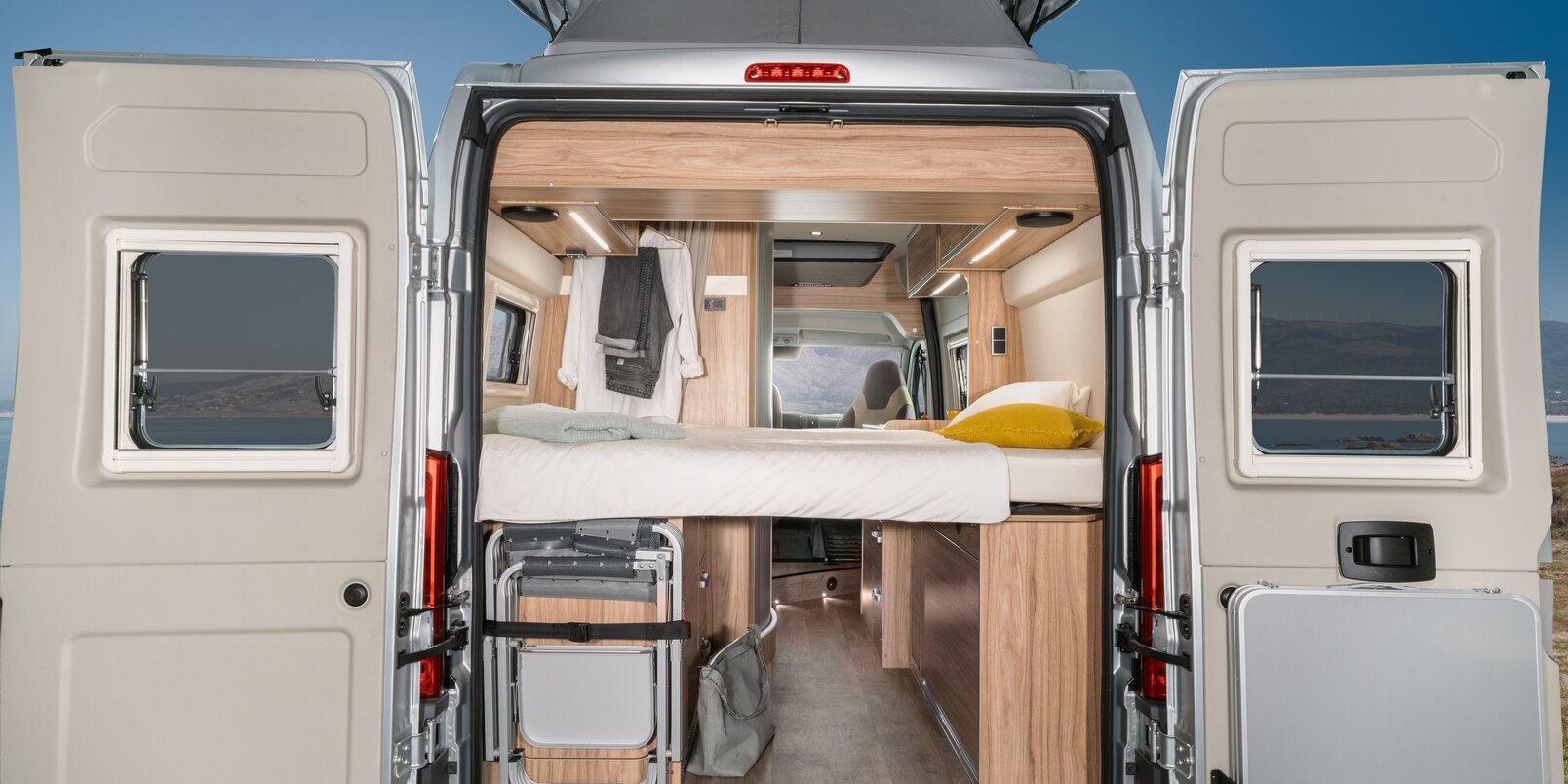 Sportello posteriore aperto, letto, sedie da campeggio e tavolo sistemati, vista all’interno fino alla cabina guida del Camper Van HYMER