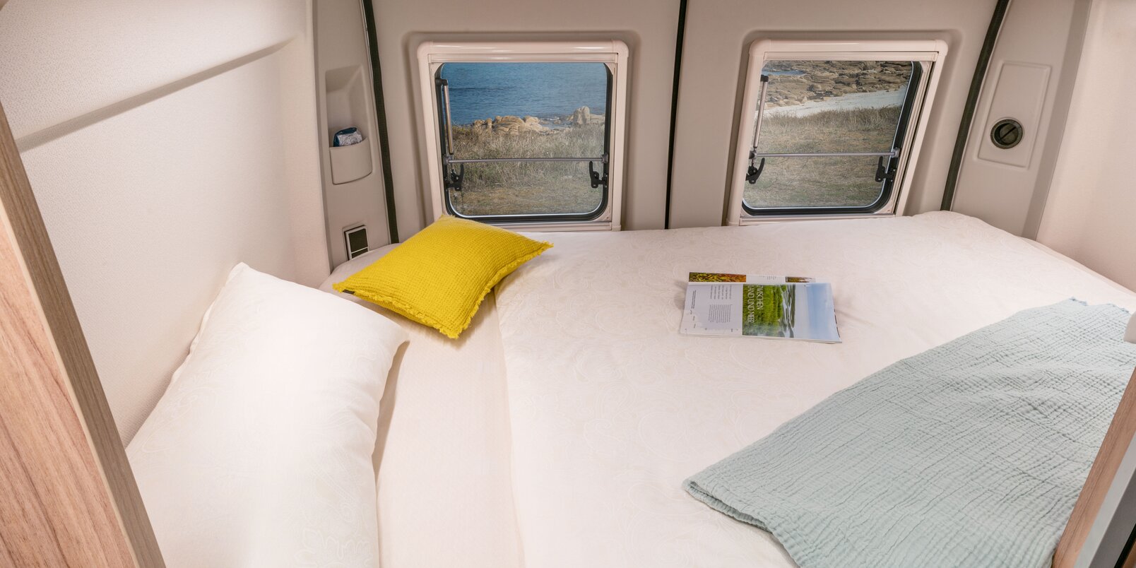 Zona notte nella parte posteriore del Camper Van HYMER: lenzuola bianche, cuscino giallo, coperta, rivista; finestra posteriore 