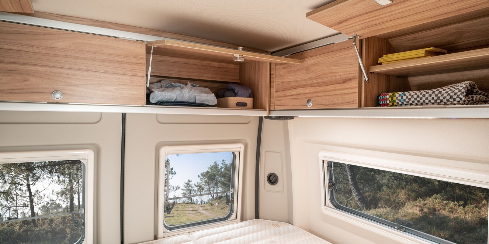 Teilweise geöffnete und befüllte Dachstauschränke über dem Bettbereich und Seitenwand-/Heckfenster im HYMER Camper Van