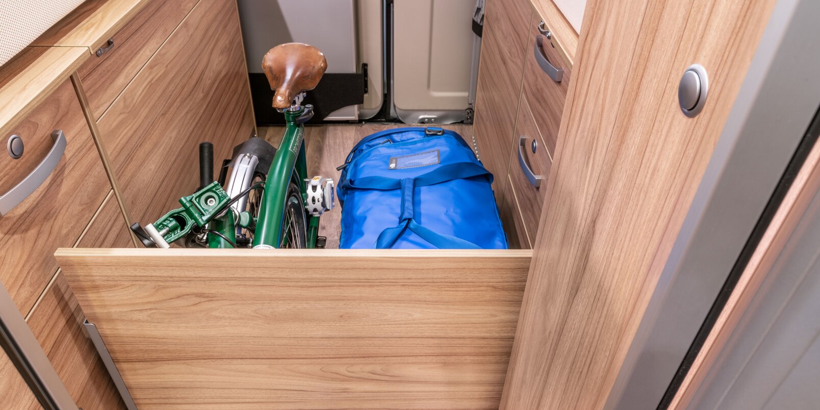 Opbergruimte gescheiden door een bord - gevuld met een blauwe reistas en groene vouwfiets - op de vloer van de HYMER-camper