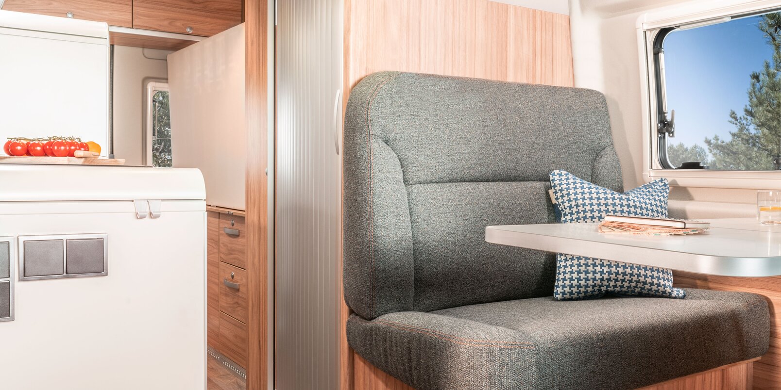 Table, living room bench with decorative cushions, window, overhead storage cupboards and refrigerator in the HYMER Fiat Camper Van