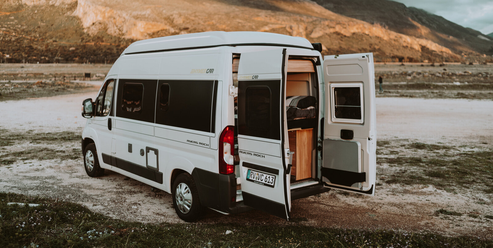 HYMER Camper Van Fiat bianco con sportelli posteriori aperti, posizionato nella sabbia, davanti ad una roccia marrone