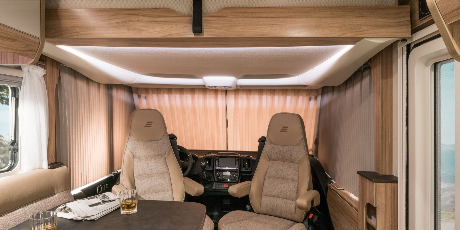 The driver's cab area darkened with a folding curtain, driver's seats, seating area and table with glasses and newspaper in the HYMER Exsis-i