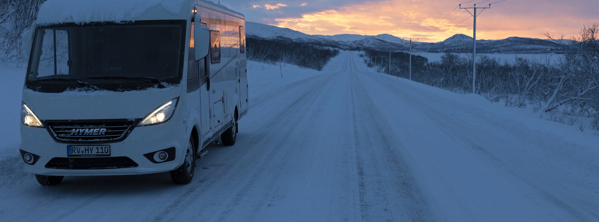 Hymermobil Exsis-i auf schneebedeckter Straße mit Stromleitung daneben und Hügeln im Hintergrund, bewölkte Abendsonne