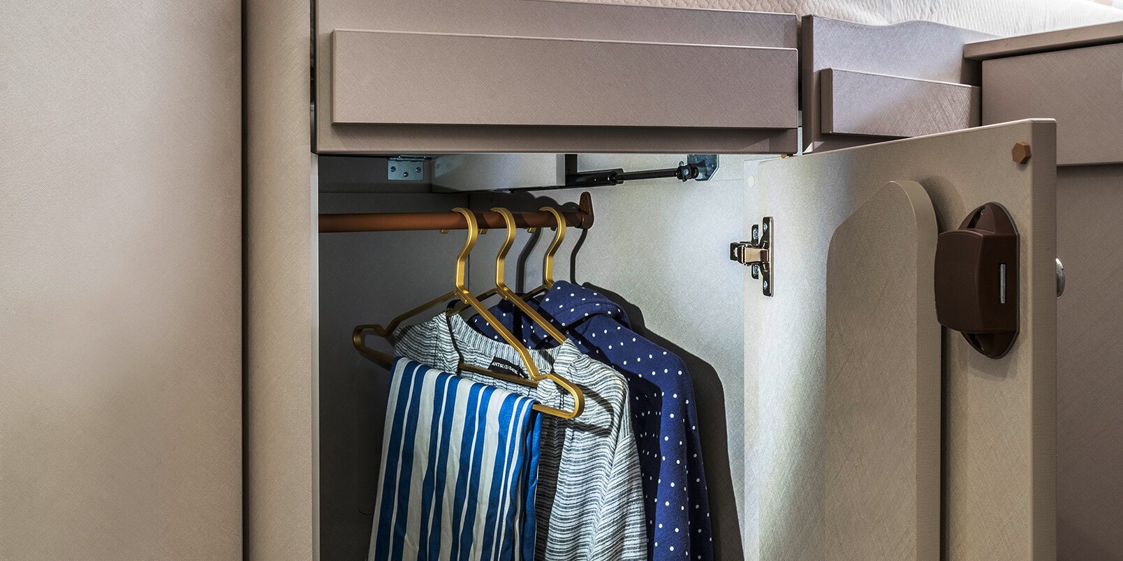 Geöffneter, beleuchteter Kleiderschrank mit behängten Kleiderbügeln auf der Stange unterm Bett im Heck des HYMER Exsis-i