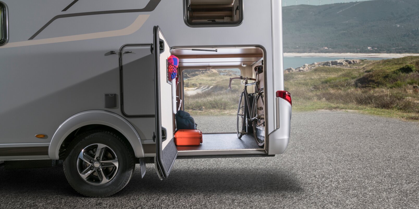 The rear garage of the HYMER Exsis-i, loaded with a red suitcase and bag, has a view of the rocky coast and the blue sky