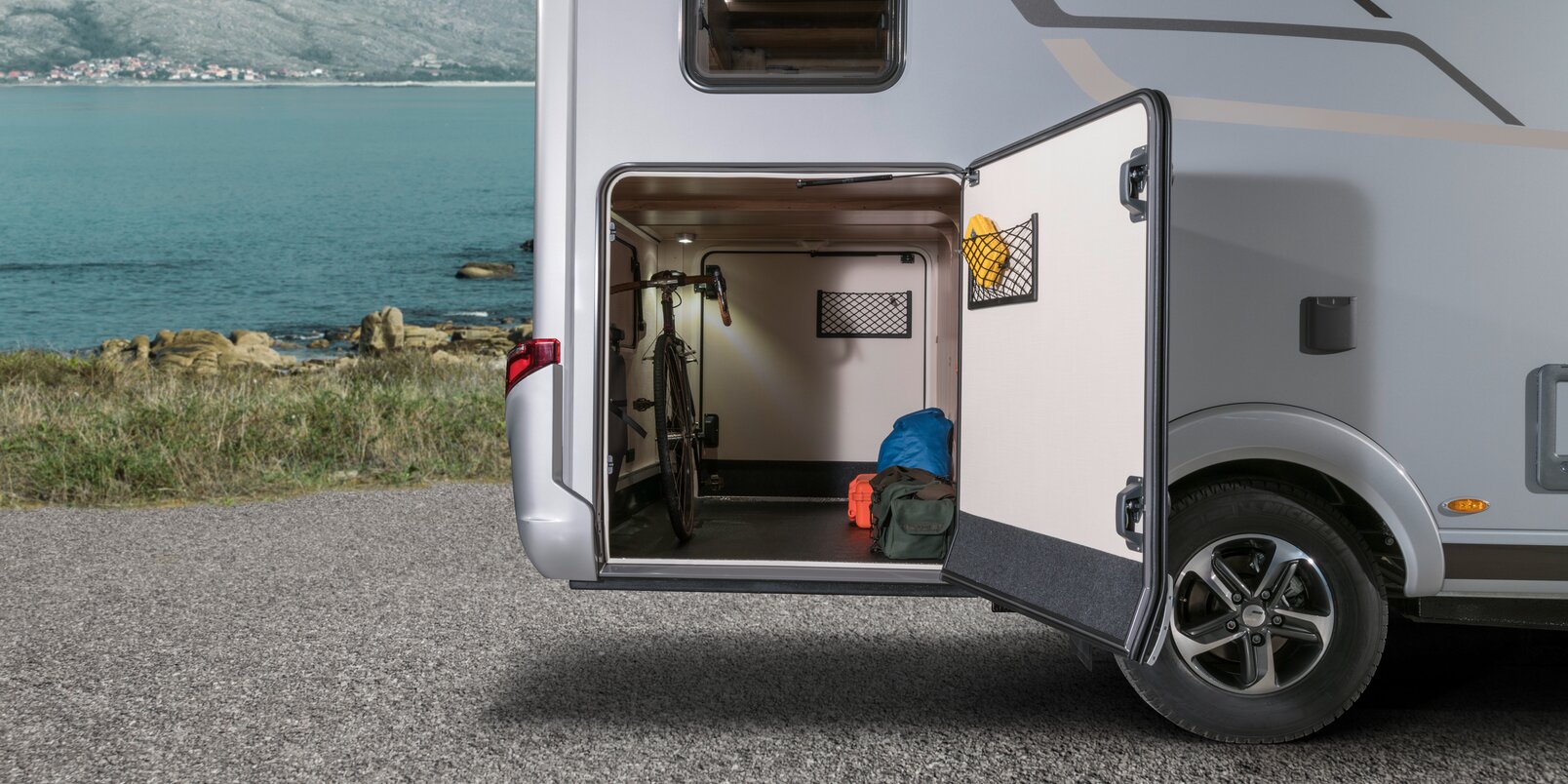 Opened door of the rear garage on the HYMER Exsis, which is loaded with luggage and bicycles in front of a rocky coast with a blue sea