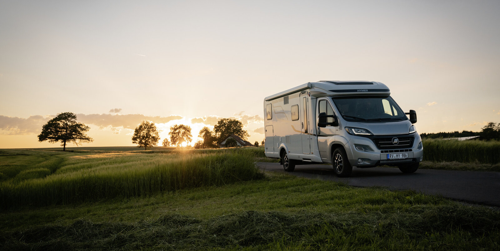 HYMER Exsis-t on a narrow street amid green meadows and cornfields at sunset
