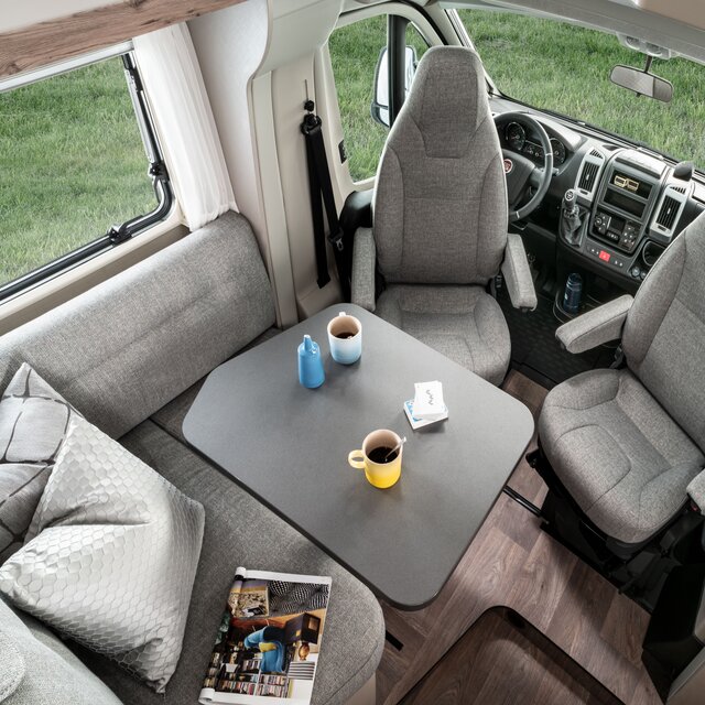 Dinette incl. sedili cabina guida nell’Exsis 580, con cuscino decorativo e rivista sulla panchina e stoviglie sul tavolo