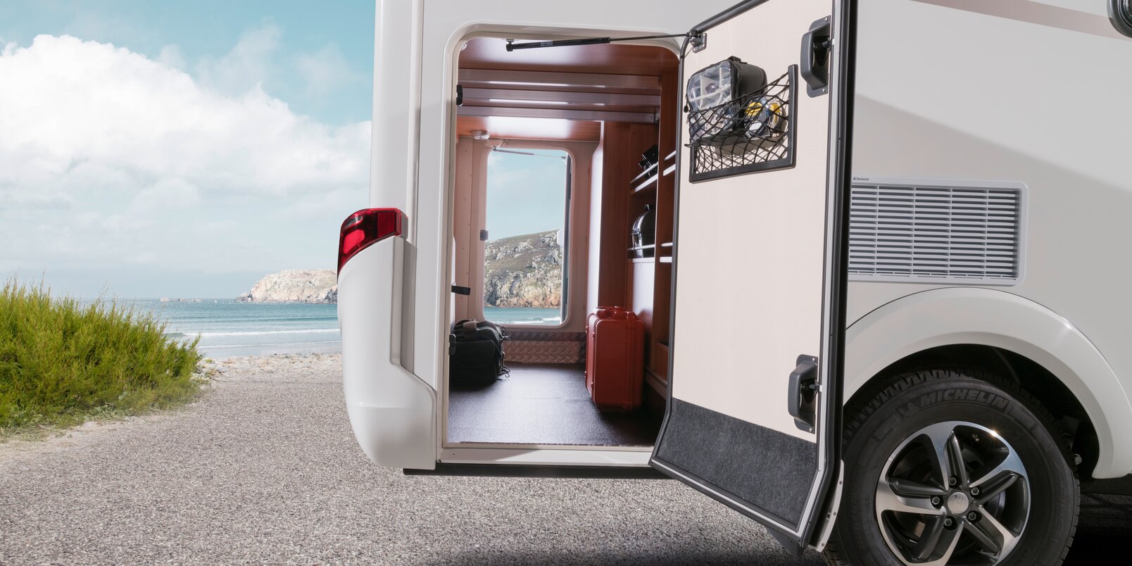 View of the rocky coast and the blue sky of the rear garage of the HYMER Exsis-t loaded with a red suitcase and bag